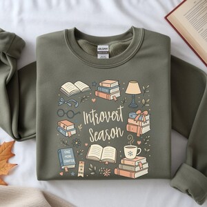 Könnte beinhalten: Olivgrünes Sweatshirt mit dem Schriftzug "Introvert Season" und Illustrationen von Büchern, Brillen, einer Lampe und einer Kaffeetasse. Das Design hat eine gemütliche, buchbezogene Ästhetik.
