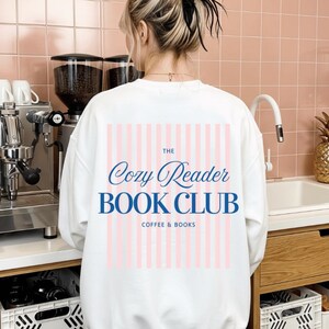 Könnte beinhalten: Ein weißes Sweatshirt mit Rundhalsausschnitt und einem Rückendruck aus vertikalen hellrosa Streifen sowie blauem Text mit der Aufschrift The Cozy Reader BOOK CLUB COFFEE & BOOKS. Das Design ist mittig auf dem langärmeligen Kleidungsstück platziert.