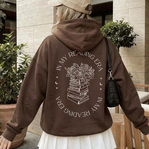 Op de afbeelding: Een donker chocoladebruine hoodie met een witte lijntekening op de achterkant. Het ontwerp toont een stapel boeken met rozen erbovenop, omringd door de tekst "IN MY READING ERA" en kleine maansikkel-symbolen.