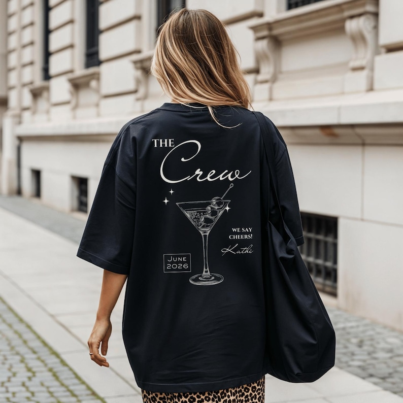 May include: A black t-shirt featuring a white graphic on the back. The design includes a martini glass, the text THE Crew, WE SAY CHEERS!, the name Kathi, and the date JUNE 2026. The shirt has short sleeves and a loose, relaxed fit.