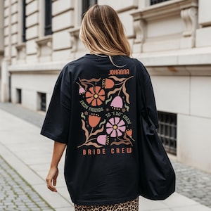 May include: Black oversized t-shirt with a floral design in orange, pink, and brown. The text "JOHANNA" is at the top, with "FROM FRIENDS TO 'I DO' CREW" and "BRIDE CREW" below the floral graphic. The shirt is worn by a person.