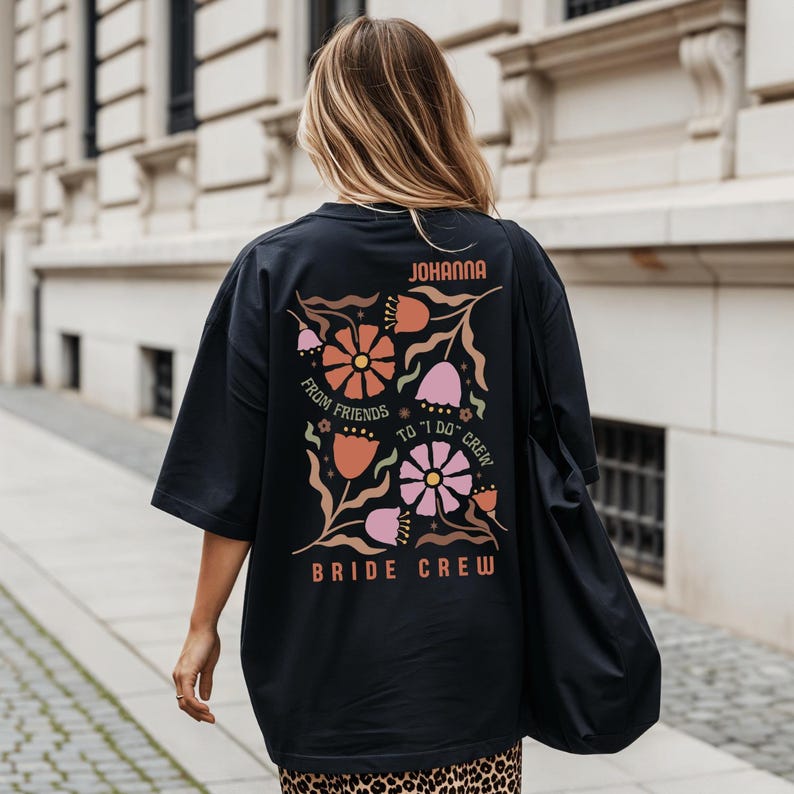 May include: Black oversized t-shirt with a floral design in orange, pink, and brown. The text "JOHANNA" is at the top, with "FROM FRIENDS TO 'I DO' CREW" and "BRIDE CREW" below the floral graphic. The shirt is worn by a person.