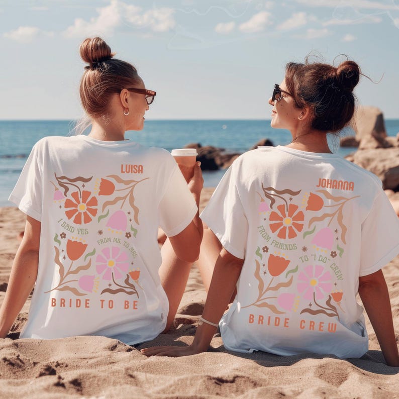 May include: White t-shirts with floral designs and text. One shirt says "Bride to Be" with "Luisa" above the design. The other says "Bride Crew" with "Johanna" above the design. The shirts are worn on a beach.