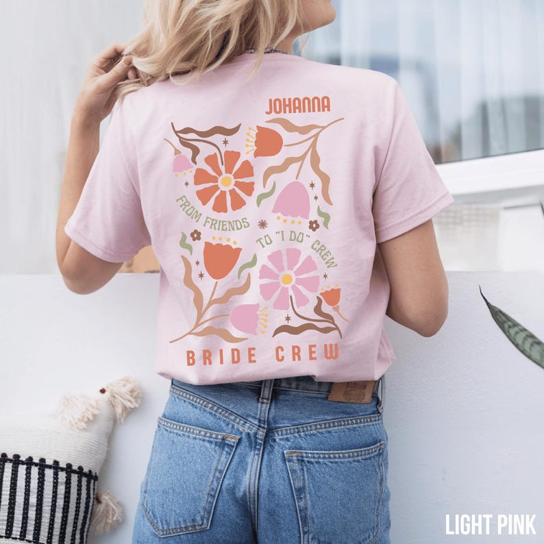 May include: Light pink t-shirt with floral design and text. The back of the shirt features a floral arrangement with the words "JOHANNA", "FROM FRIENDS TO 'I DO' CREW", and "BRIDE CREW". The shirt is made of a soft material.