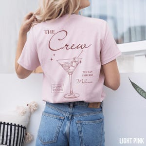 May include: A light pink short-sleeve t-shirt featuring a dark red graphic of a martini glass with olives. The text reads THE Crew, WE SAY CHEERS!, Melanie, and JUNE 2026. The design is printed on the back of the shirt, which is worn with blue jeans.