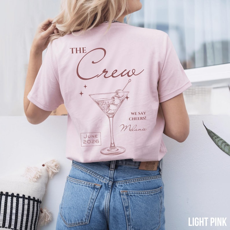 May include: A light pink short-sleeve t-shirt featuring a dark red graphic of a martini glass with olives. The text reads THE Crew, WE SAY CHEERS!, Melanie, and JUNE 2026. The design is printed on the back of the shirt, which is worn with blue jeans.