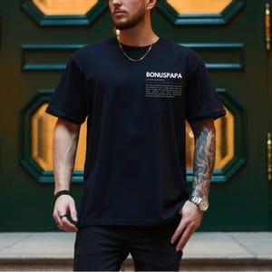 May include: A black t-shirt with a white print that reads "BONUSPAPA" and a definition of the word. The shirt is being worn by a person with tattoos on their arm.