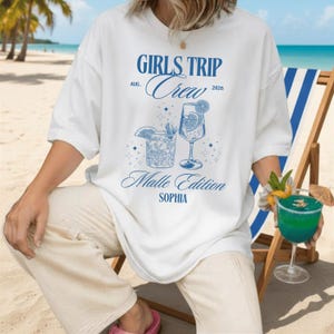 Puede incluir: Camiseta blanca con un dise&ntilde;o azul que dice "Girls Trip Crew" con "Aug. 2026" y "Malle Edition SOPHIA". El dise&ntilde;o incluye ilustraciones de c&oacute;cteles. La persona est&aacute; sentada en la playa.