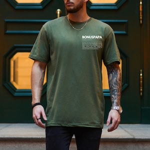 May include: A person wearing a green t-shirt with the text "BONUSPAPA" and a definition of the word. The person is wearing black pants and has a tattoo on their arm.