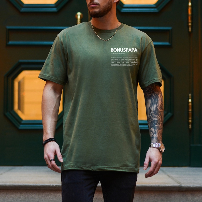 May include: A person wearing a green t-shirt with the text "BONUSPAPA" and a definition of the word. The person is wearing black pants and has a tattoo on their arm.