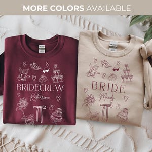 May include: Two crewneck sweatshirts, one burgundy and one beige, with white graphic designs. The burgundy sweatshirt says "BRIDE CREW" and the beige one says "BRIDE". Both have wedding-themed illustrations. The text "MORE COLORS AVAILABLE" is at the top.
