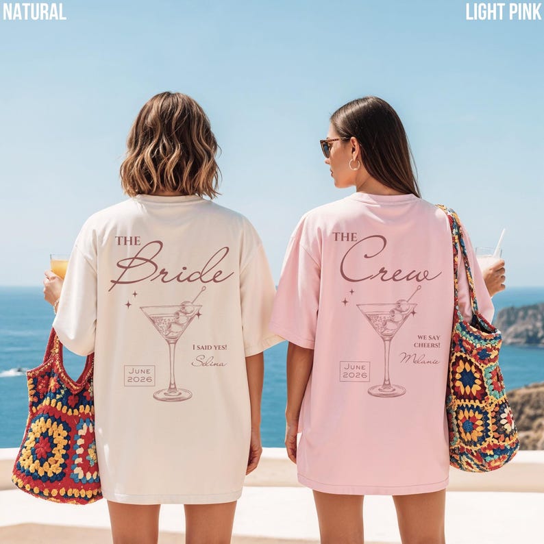 May include: Two short-sleeve t-shirts, one natural and one light pink, each featuring a martini glass graphic, star accents, and text. The natural shirt reads THE Bride, I SAID YES!, JUNE 2026, and Selina. The pink shirt reads THE Crew, WE SAY CHEERS!, JUNE 2026, and Melanie.