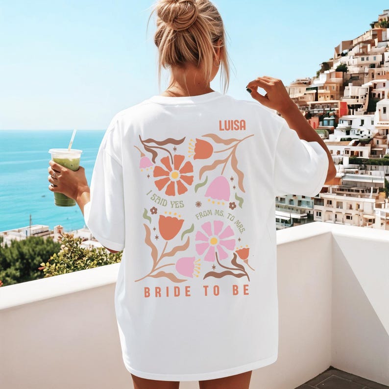May include: White t-shirt with a floral design in orange, pink, and brown. The text on the back reads "LUISA", "I SAID YES", "FROM MS. TO MRS.", and "BRIDE TO BE". The shirt is a relaxed fit.