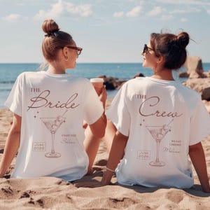 May include: Two white t-shirts featuring a brown graphic of a martini glass with olives. The left shirt displays the text THE Bride, I SAID YES!, Selina, and JUNE 2026. The right shirt displays the text THE Crew, WE SAY CHEERS!, Melanie, and JUNE 2026.
