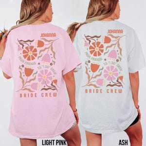 May include: Light pink and ash-colored t-shirts with floral designs and text. The shirts feature the name "JOHANNA" at the top, with "FROM FRIENDS TO 'I DO' CREW" and "BRIDE CREW" below. The floral design includes orange, pink, and beige elements.