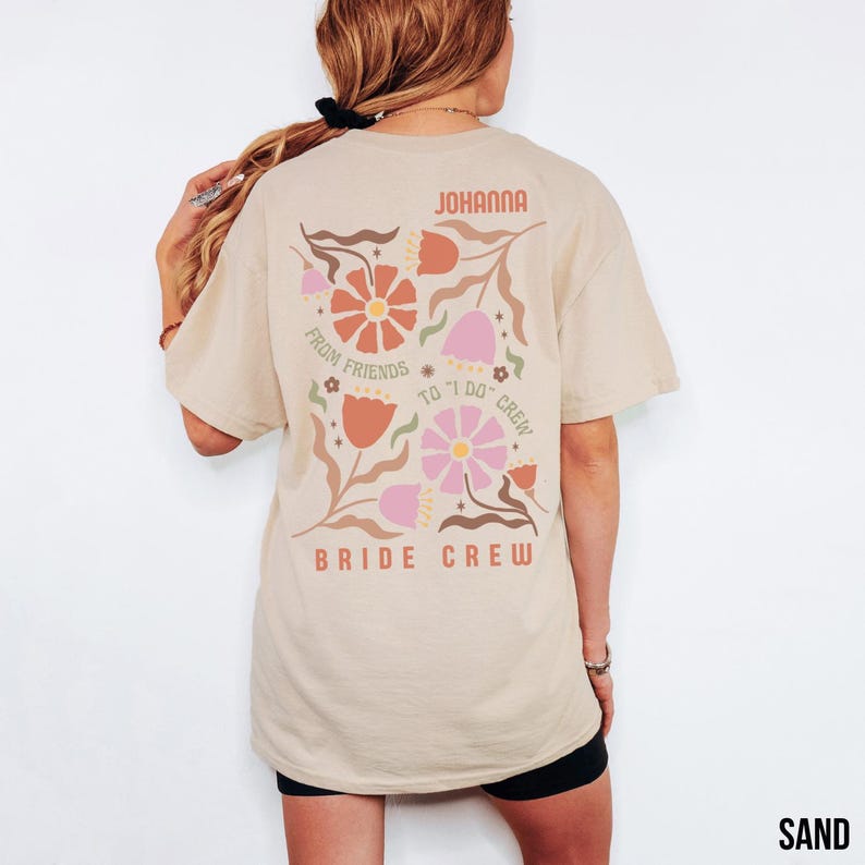 May include: Sand-colored t-shirt with a floral design on the back. The design includes flowers in shades of orange, pink, and brown, with the words "JOHANNA", "FROM FRIENDS TO I DO CREW", and "BRIDE CREW".