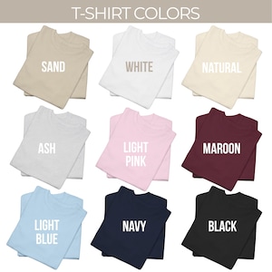 May include: A grid of nine folded t-shirts in various colors labeled with text: Sand, White, Natural, Ash, Light Pink, Maroon, Light Blue, Navy, and Black. The header text reads T-SHIRT COLORS. Each shirt is displayed in a solid, flat color.
