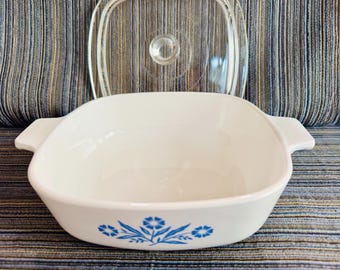 May include: White square casserole dish with blue floral design and clear glass lid. The dish has two handles and is made of ceramic. A classic piece of kitchenware.