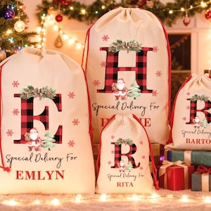 May include: Four beige Christmas gift bags with red drawstrings. Each bag has a large letter in a red and black plaid pattern, a Santa Claus illustration, and the words "Special Delivery For" and a name.