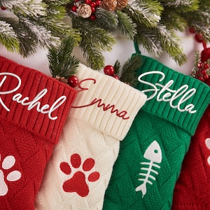 May include: Four personalised Christmas stockings in red, cream, and green, with names embroidered in white and red. The red stockings feature white paw prints, and the green stocking has a fishbone design. Festive holiday decor.