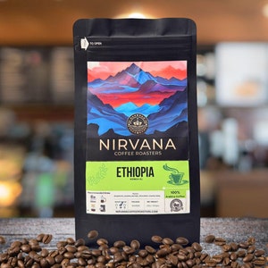 May include: A black bag of Nirvana Coffee Roasters Ethiopia Yirgacheffe coffee beans. The bag features a colorful mountain landscape design with the text "Nirvana Coffee Roasters" and "Ethiopia" in white. The bag also includes a green leaf design and the text "100% Arabica Coffee".