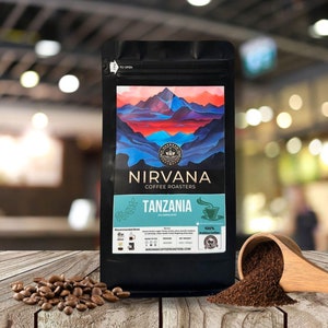 May include: A black bag of Nirvana Coffee Roasters Tanzania Kolmeruard coffee beans. The bag features a colorful mountain landscape design. The bag is sitting on a wooden table with a scoop of coffee beans next to it.