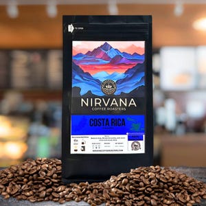May include: A black bag of Nirvana Coffee Roasters Costa Rica coffee beans. The bag features a mountain landscape design in blue, pink, and orange. Coffee beans are scattered around the bag on a wooden surface.