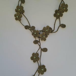 Può includere: Una collana fatta a mano all'uncinetto con un design floreale. La collana è verde oliva con piccole perline verdi rotonde al centro di ogni fiore. I fiori sono collegati da una struttura intrecciata a forma di vite.