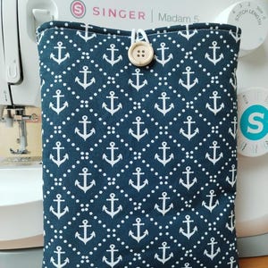 May include: A navy blue book sleeve with a white anchor pattern and a wooden button fastening. The sleeve is designed to protect books and features a nautical theme. The Singer sewing machine is in the background.