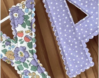 May include: A fabric letter Y featuring a floral pattern with purple flowers, green leaves, and orange buds, alongside a fabric letter P in light purple with a white star pattern. Both letters have scalloped edges and are attached to a white cord.
