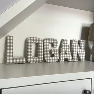 May include: The name "LOGAN" is spelled out in large, fabric-covered letters. The letters are in a neutral color palette, with a gray and white gingham pattern. The letters are displayed on a white shelf, creating a decorative element for a nursery or child's room.