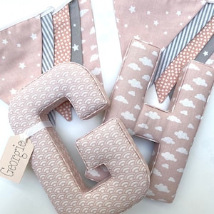 Puede incluir: Letras de tela rosa que deletrean el nombre "Georgie" con un diseño estampado rosa y blanco. Las letras están rodeadas de una guirnalda rosa y blanca con estrellas, corazones y nubes.