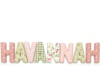 May include: Fabric-covered letters spelling the name HAVANNAH. The letters feature various patterns including pink with white polka dots, green and white gingham, pink stripes, and a floral print with small red flowers and green leaves on a cream background.