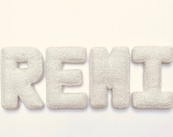 May include: White felt letters spelling out the name "REMI" on a white background. The letters are fluffy and have a slightly textured surface. The letters are arranged in a straight line, with the first letter "R" on the left and the last letter "I" on the right. The letters are slightly spaced apart. The text "@laurasloveletters" is written in black below the letters.