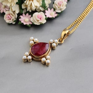 Peut inclure: Un collier avec un pendentif en or orné d'une pierre de rubis en forme de larme entourée de perles blanches. Le collier a une chaîne délicate.