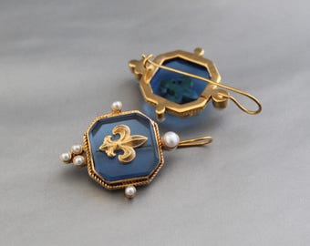 May include: Two gold-toned brooches featuring an octagonal blue glass center with a gold fleur-de-lis emblem. Each piece is accented with small white pearls along the edges and includes a thin gold pin mechanism on the back for attachment.