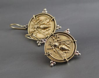Op de afbeelding: Een paar gouden oorbellen met een bijendesign. De oorbellen zijn gemaakt van metaal en hebben een ronde vorm. De bij bevindt zich in het midden van de oorbel en is omgeven door een cirkel van kleine kralen.