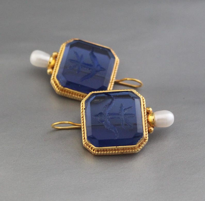 May include: A pair of gold earrings with blue glass square-shaped stones. Each stone has an etched design of an anchor. The earrings have a pearl accent on the bottom.