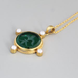 Op de afbeelding: Een goudkleurige hangerketting met een groene, ovaalvormige steen met een geëtst ontwerp. De hanger is versierd met vier witte parels en hangt aan een fijne gouden ketting. De ketting wordt gepresenteerd tegen een lichtgrijze achtergrond.