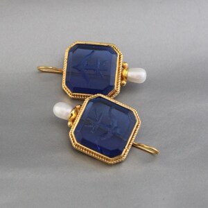May include: A pair of gold earrings with blue glass square-shaped stones and a pearl accent. The stones have a raised design of an anchor.