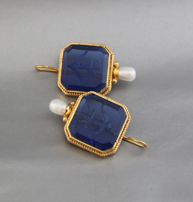 May include: A pair of gold earrings with blue glass square-shaped stones and a pearl accent. The stones have a raised design of an anchor.