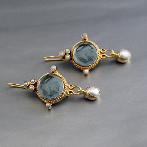 K&ouml;nnte beinhalten: Ein Paar goldene Ohrringe mit blauen Edelstein- und Perlenakzenten. Die Ohrringe haben einen runden, facettierten blauen Edelstein, der von einem goldenen Rahmen und kleinen Perlen umgeben ist. Jeder Ohrring hat eine einzelne Perle, die an der Unterseite h&auml;ngt.