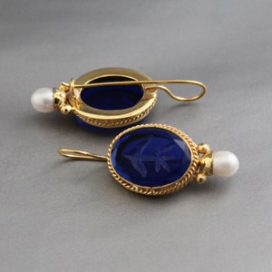 Puede incluir: Un par de pendientes de oro con piedras ovaladas de vidrio azul y un solo acento de perla en cada pendiente.