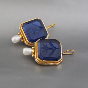 May include: A pair of gold earrings with blue glass square-shaped stones. Each stone has a gold rim and a small pearl attached to the top.