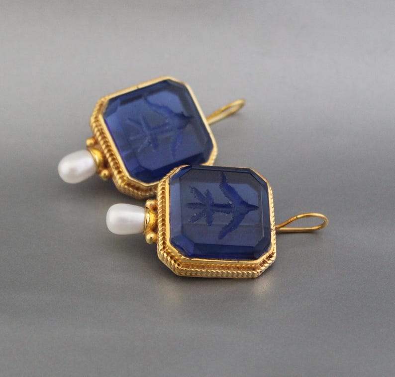 May include: A pair of gold earrings with blue glass square-shaped stones. Each stone has a gold rim and a small pearl attached to the top.