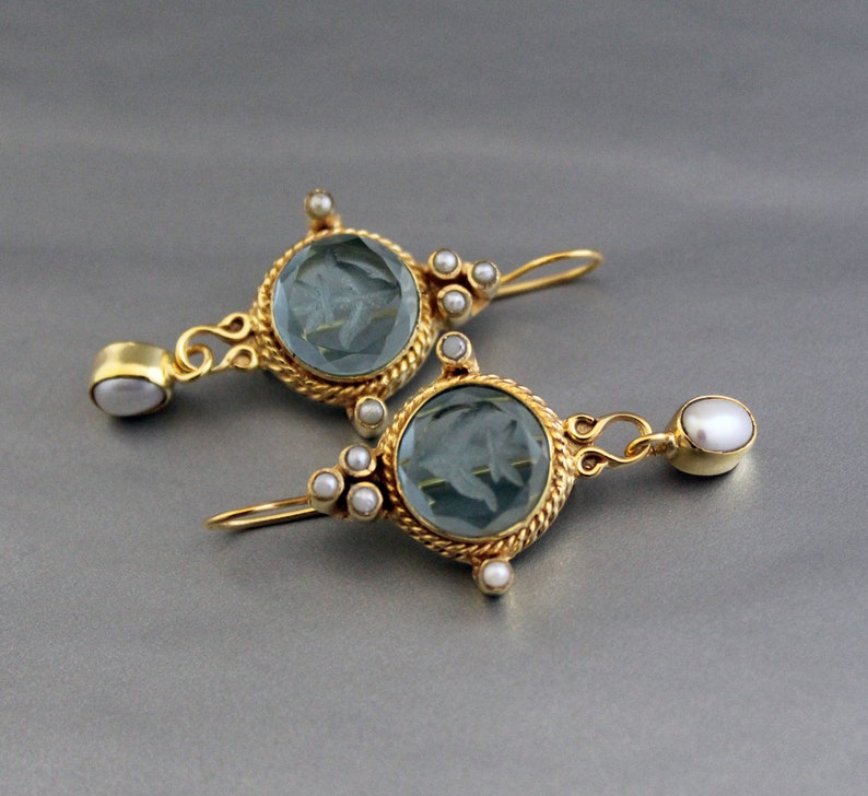 K&ouml;nnte beinhalten: Ein Paar goldene Ohrringe mit blauen Glassteinen und wei&szlig;en Perlenakzenten. Die Ohrringe haben ein detailliertes Design mit einem seilartigen Rand um die Steine.