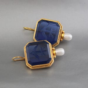 May include: A pair of gold earrings with blue glass insets and a pearl dangle. The glass insets feature an etched design of a bird.