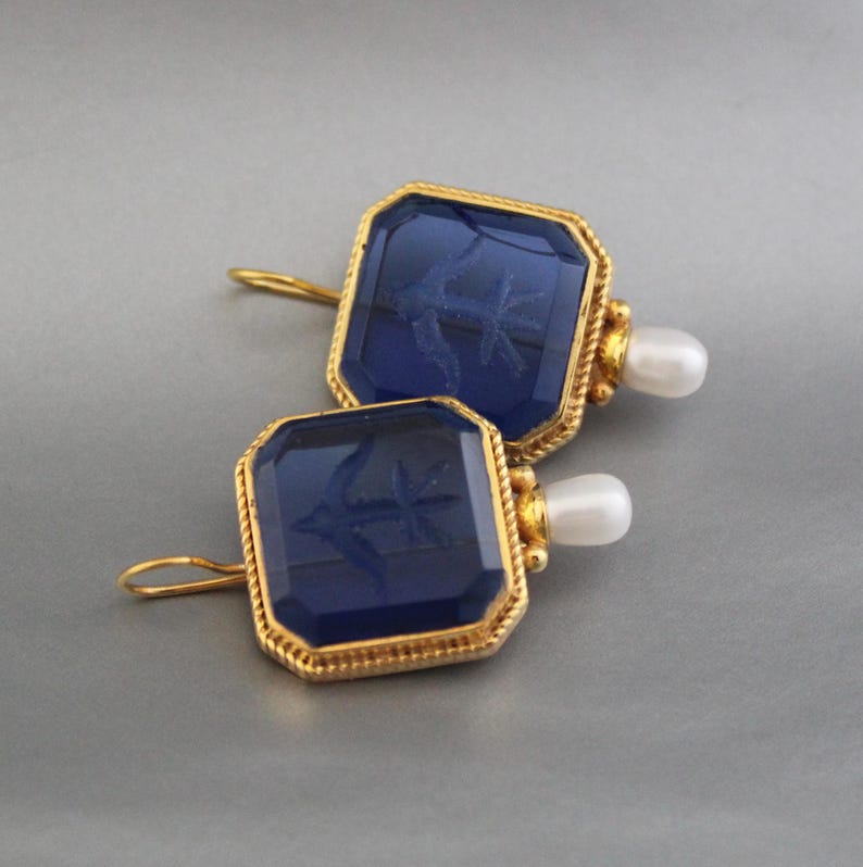 May include: A pair of gold earrings with blue glass insets and a pearl dangle. The glass insets feature an etched design of a bird.