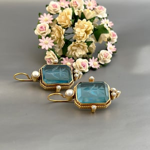Puede incluir: Un par de pendientes de oro con piedras de cristal azul y detalles de perlas. Las piedras son rectangulares y tienen un acabado mate. Los pendientes son de estilo colgante.