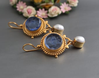 Op de afbeelding: Een paar gouden oorbellen met blauwe glazen cabochons en witte parel accenten. De oorbellen hebben een gedetailleerd ontwerp met een ronde vorm en een druppel stijl.